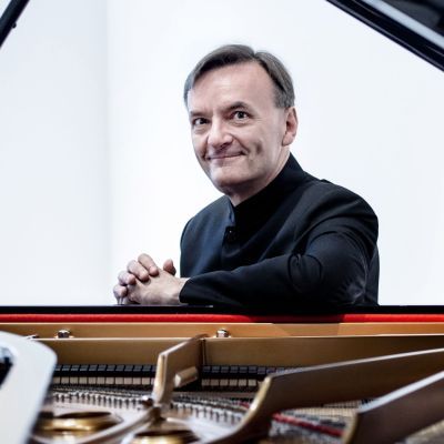 A smiling Stephen Hough wearing a black suit sat behind a piano with a white backdrop